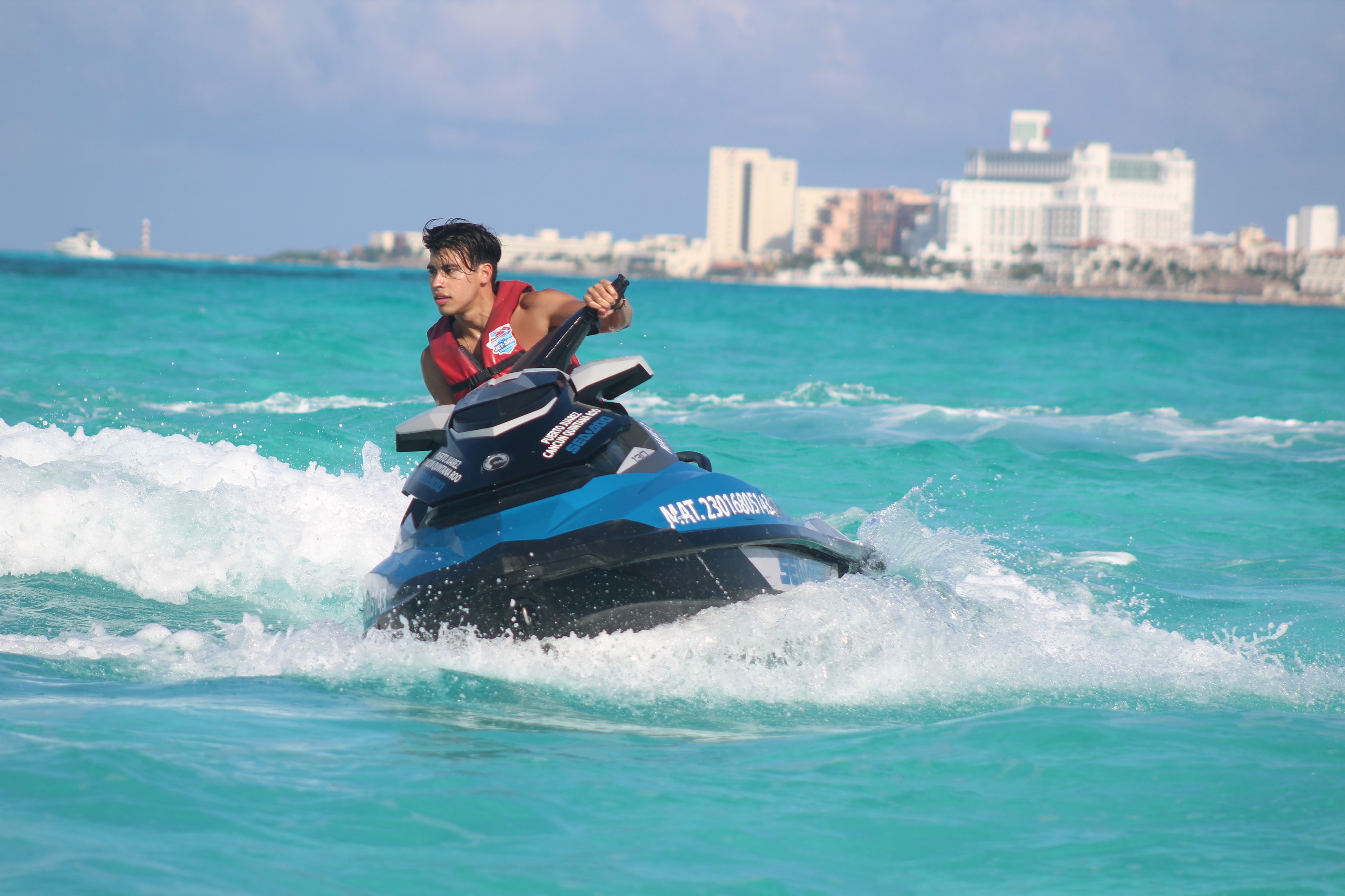 Young man riding a jet ski through choppy turquoise water with Cancun hotels in the background