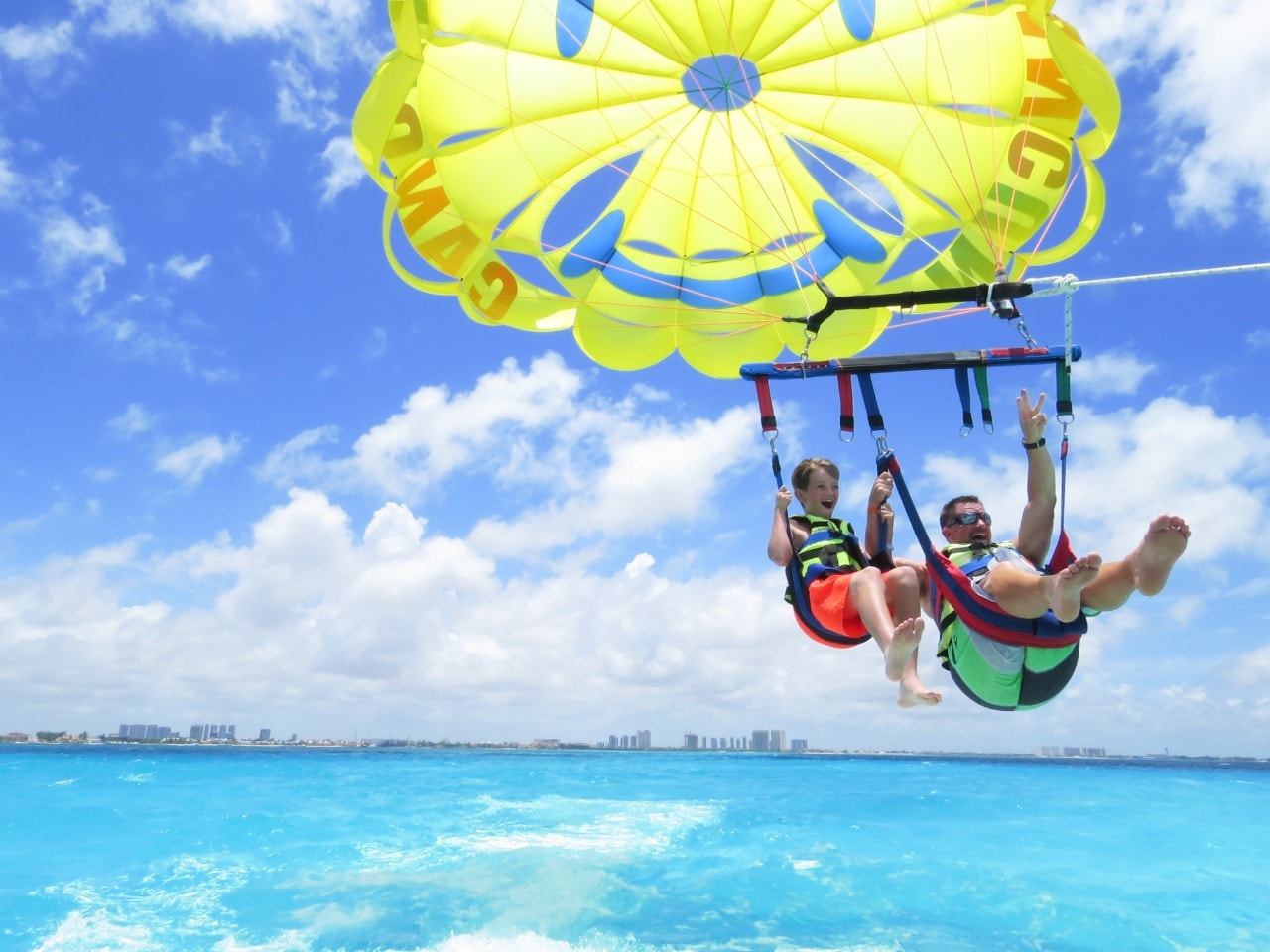 Father and son waving and smiling while parasailing high above the Caribbean with Cancun skyline behind
