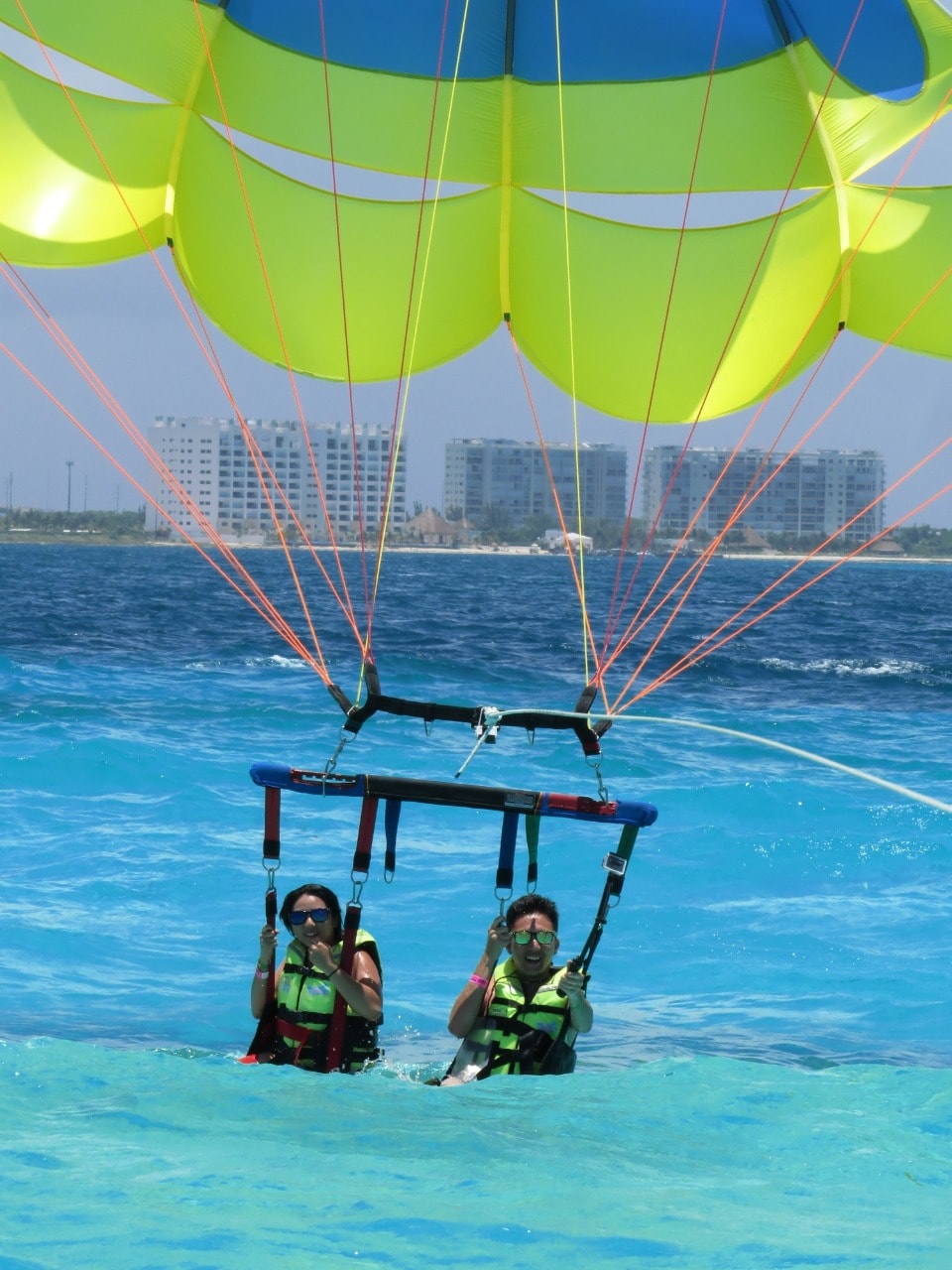 Couple touching down into the turquoise Caribbean water under a lime-green parasail with Cancun hotels behind