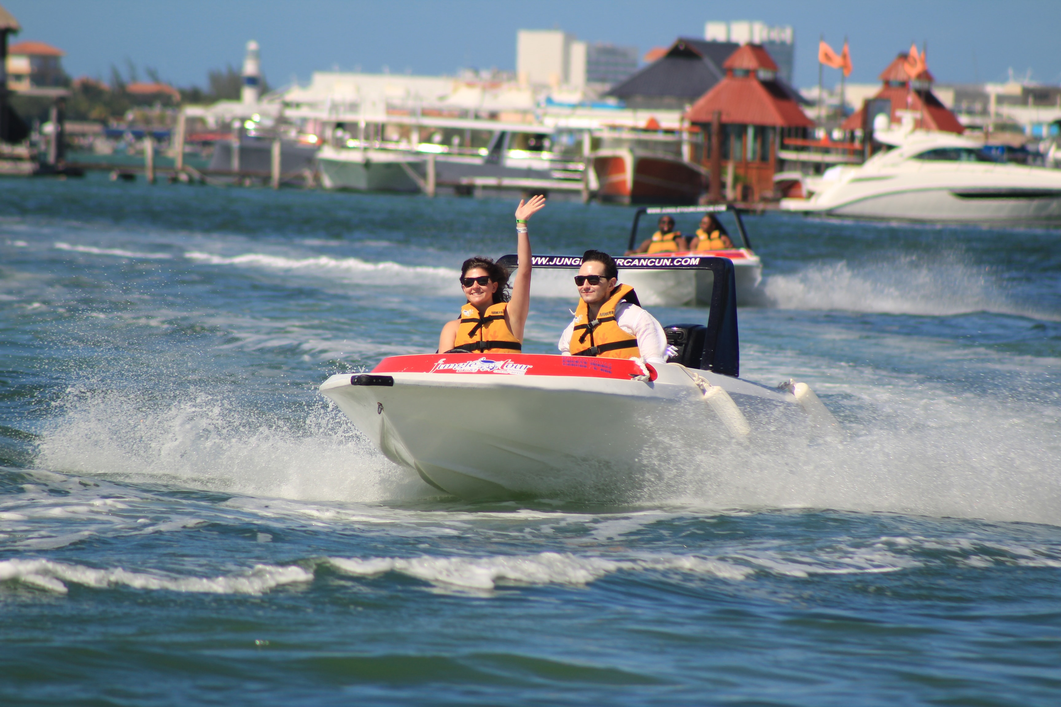 Smiling couple in life jackets waving from a Jungle Tour speedboat on the Cancun lagoon