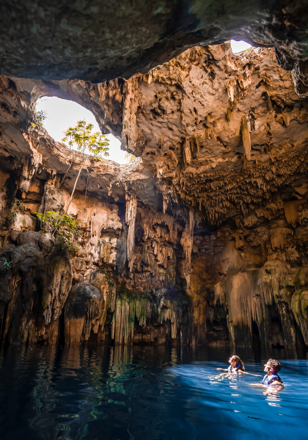 Two swimmers inside a dramatic cenote cave with stalactites and natural light beams