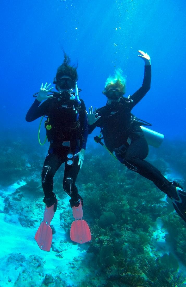Two scuba divers waving and posing playfully above a vibrant coral reef in the clear blue waters of Cozumel